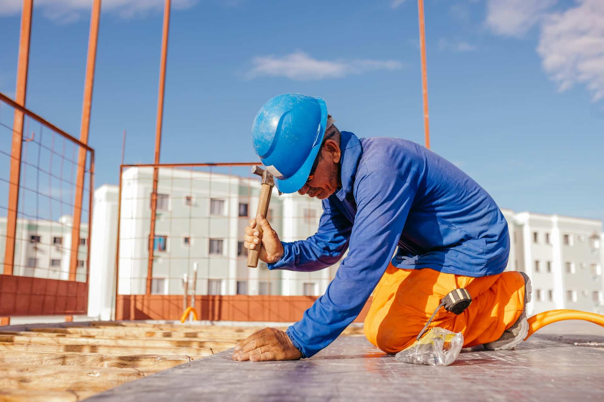 Construction worker in a blue hard hat and high-vis overalls kneeling to hammer a nail on a building site